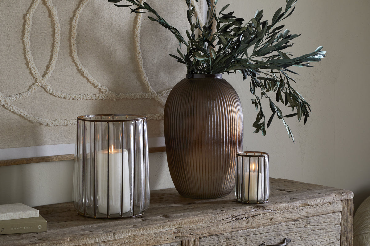 A rustic wooden console displays a large ribbed brown vase with olive branches, antique brass tealight holders, and the nkuku Hava Lantern - Clear with a lit pillar candle. Textured beige wall art hangs in the background.