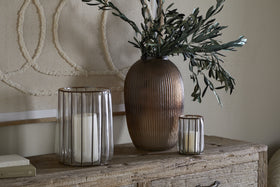 A rustic wooden sideboard displays a ribbed brown vase with leafy stems, flanked by two nkuku Hava Lanterns - Clear, one large and one small, each holding a white pillar candle. Above hangs textured abstract wall art.