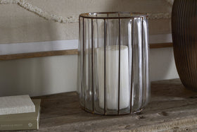 The Hava Lantern - Clear by nkuku features a large white candle inside a ribbed glass hurricane with antique brass accents, displayed on rustic wood beside stacked books against a neutral textured background.