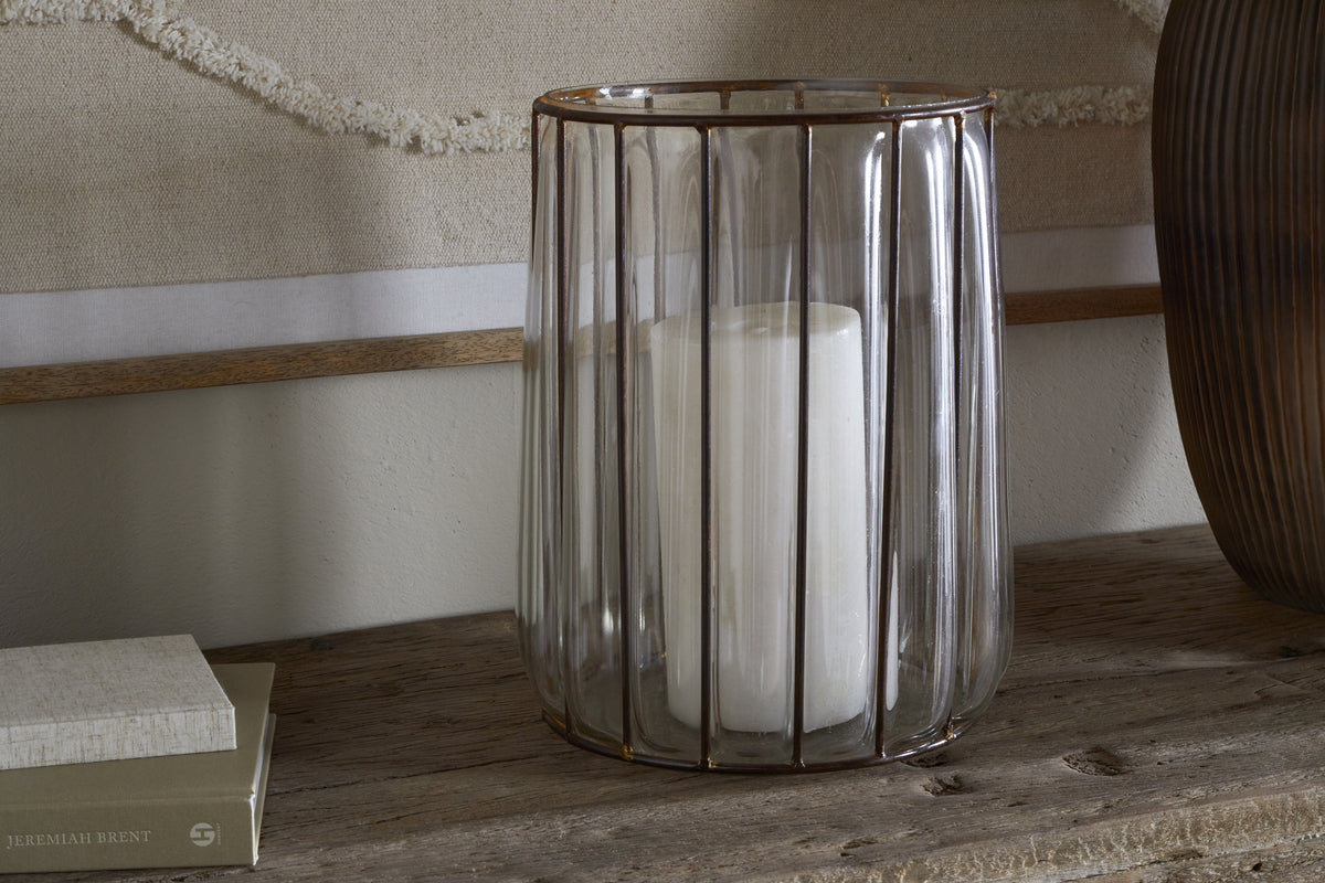 The Hava Lantern - Clear by nkuku features a large white candle inside a ribbed glass hurricane with antique brass accents, displayed on rustic wood beside stacked books against a neutral textured background.