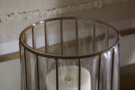 A close-up of the nkuku Hava Lantern - Clear shows its cylindrical glass body with vertical antique brass bars in an openwork design. A white pillar candle glows inside, while blurred textured fabric and a wooden surface form the background.