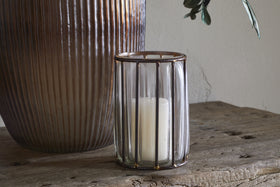 The nkuku Hava Lantern - Clear features vertical metal bars and antique brass accents, holding a white pillar candle. It rests on a rustic wooden surface next to a large ribbed ceramic vase with a plant leaf against a neutral wall.