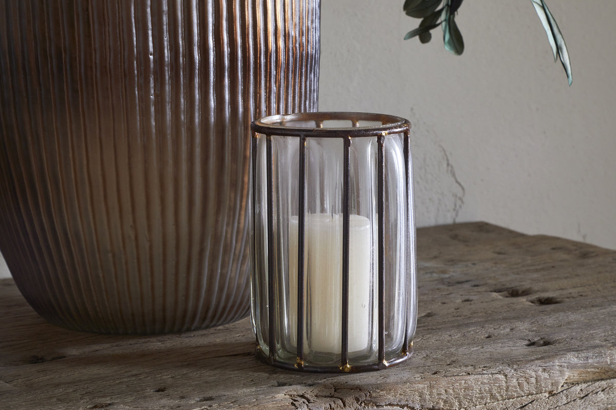 The nkuku Hava Lantern - Clear features vertical metal bars and antique brass accents, holding a white pillar candle. It rests on a rustic wooden surface next to a large ribbed ceramic vase with a plant leaf against a neutral wall.
