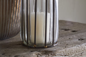 The Hava Lantern - Clear by nkuku features a white candle in a clear glass holder with vertical metal bars and openwork design, set on rustic wood. A ribbed metallic accent appears on the left, and soft lighting adds a cozy touch.