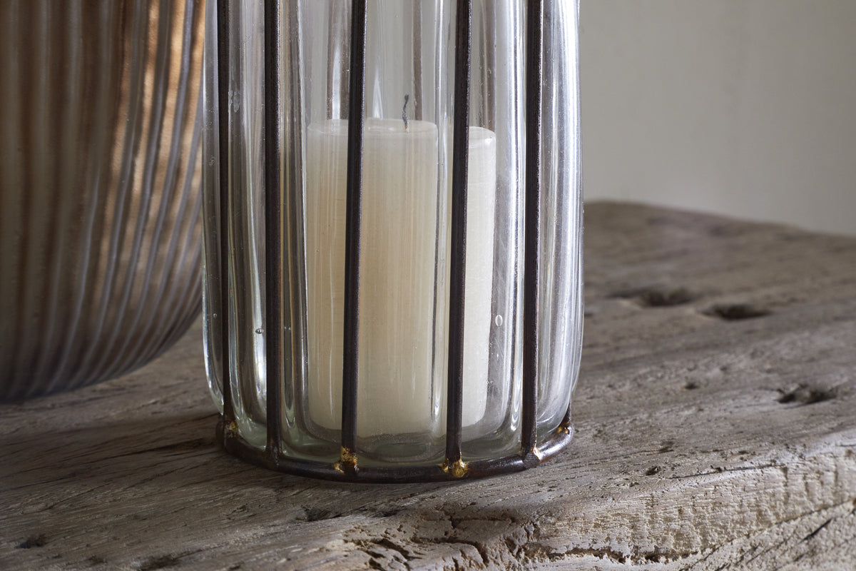 The Hava Lantern - Clear by nkuku features a white candle in a clear glass holder with vertical metal bars and openwork design, set on rustic wood. A ribbed metallic accent appears on the left, and soft lighting adds a cozy touch.