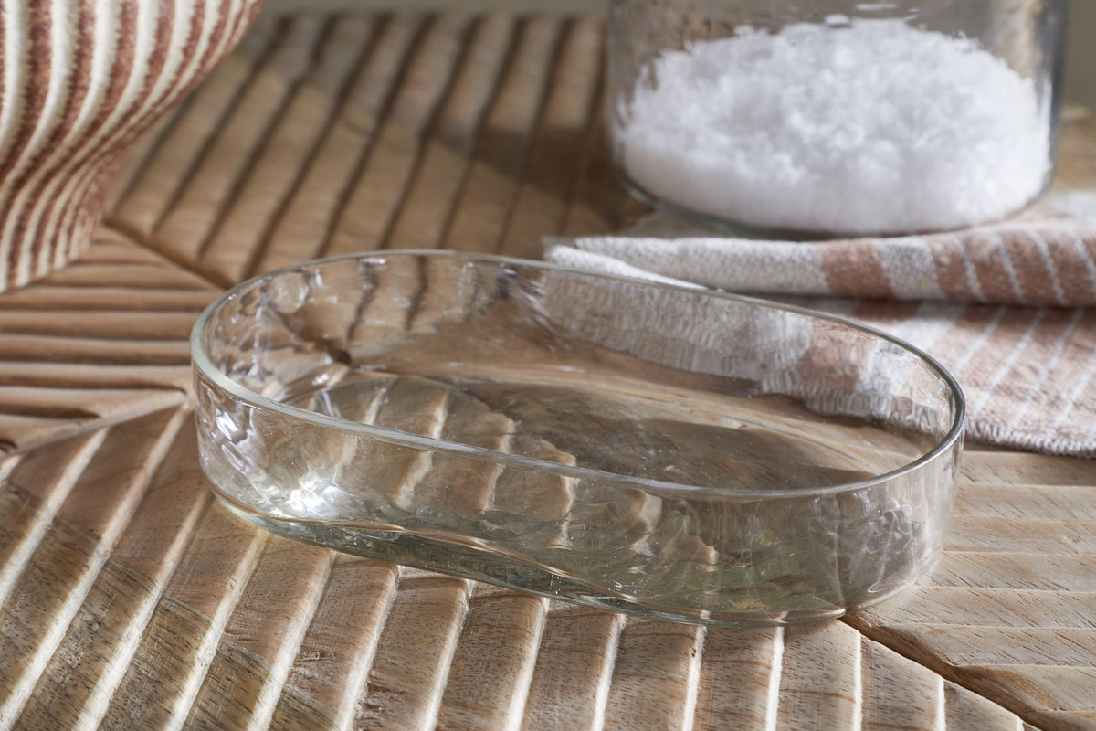 The nkuku Ilcoso Recycled Hammered Glass Soap Dish - Clear rests on a ribbed wooden surface, accompanied by a jar of flaked salt and a folded white and tan striped cloth, creating a serene bathroom scene in soft natural light.