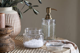 The nkuku Ilcoso Recycled Hammered Glass Storage Pot - Clear filled with white bath salts sits on a wooden surface next to its lid, accompanied by a glass soap dispenser, green leaves in a textured vase, and a brown candle in a wooden holder.