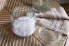 The Ilcoso Recycled Hammered Glass Storage Pot by nkuku, filled with coarse white salt, sits open on a wooden table beside its lid, a striped beige and white cloth, and a glass container of clear liquid in natural light.