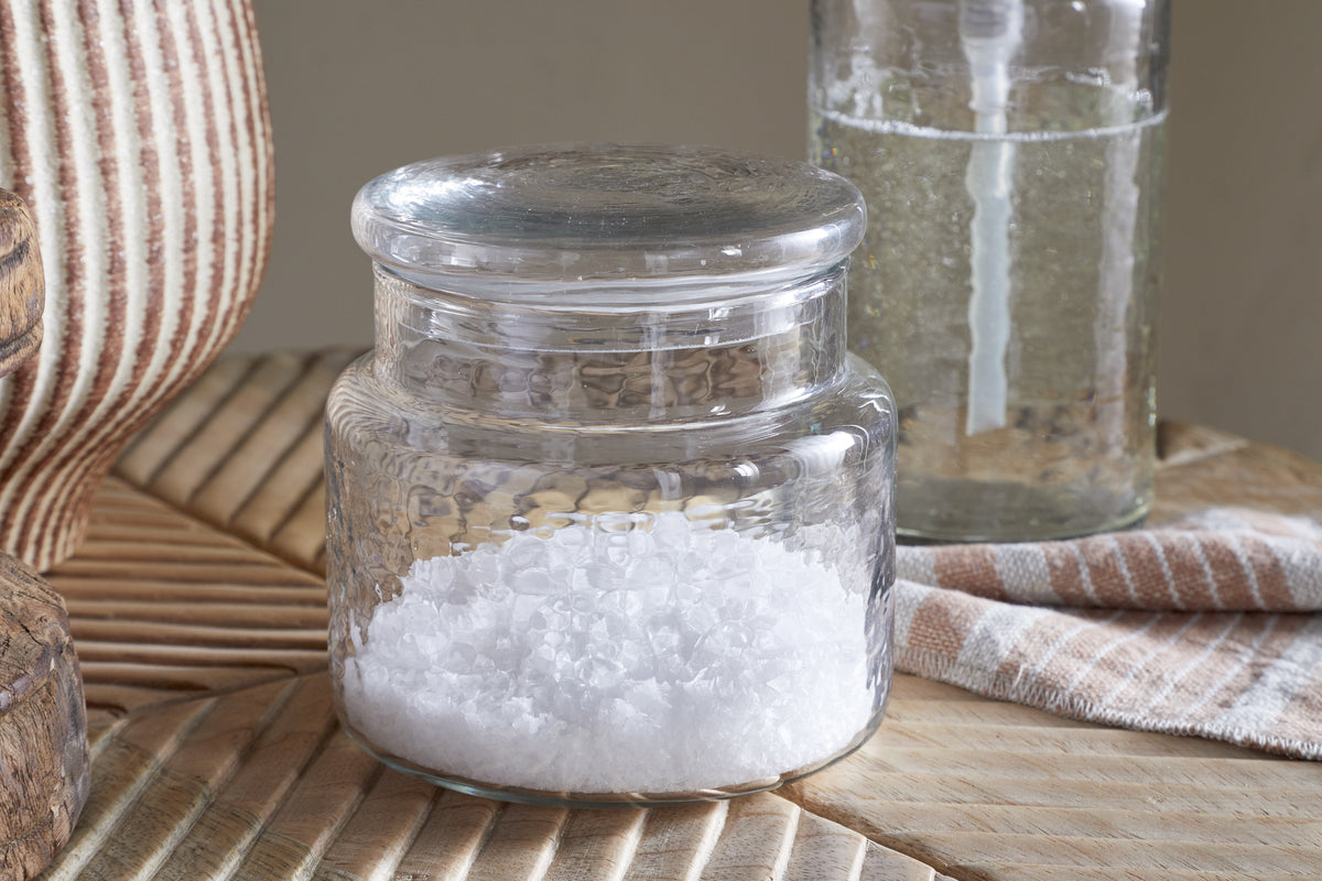The Ilcoso Recycled Hammered Glass Storage Pot by nkuku, filled with coarse white sea salt, sits on a patterned light wooden surface. In the background are a glass bottle, striped cloth, and part of a wicker or wooden object.