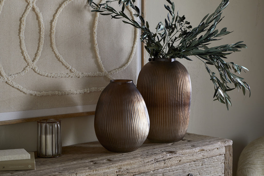 The Linsy Ribbed Glass Vase in Smoke Brown by nkuku, paired with a shorter clear vase filled with green branches, sits on a rustic wooden table alongside a candle holder, book, and textured wall art for minimalist, earthy decor.
