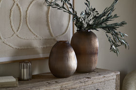 The Linsy Ribbed Glass Vase in Smoke Brown by nkuku, paired with a shorter clear vase filled with green branches, sits on a rustic wooden table alongside a candle holder, book, and textured wall art for minimalist, earthy decor.