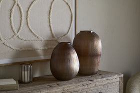 The nkuku Linsy Ribbed Glass Vase in Smoke Brown sits on a rustic wooden table by a candle and closed book, with neutral textile wall art featuring three large textured circles on a beige background behind it.