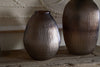 Two Linsy Ribbed Glass Vases by nkuku in smoke brown—one oval, one cylindrical—rest on a rustic wooden surface before textured beige art, creating a warm, earthy, and elegant look.