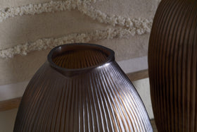 Close-up of the nkuku Linsy Ribbed Glass Vase in Smoke Brown, featuring an oval shape and asymmetrical opening. Warm light highlights its grooves as it stands before cream-textured fabric, with a similar piece partially visible on the right.