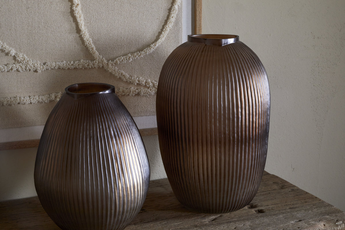 Two Linsy Ribbed Glass Vases by nkuku, in smoke brown and varying sizes with a semi-opaque finish, rest on a rustic wooden surface. Behind them hangs textured neutral artwork on a smooth beige wall.