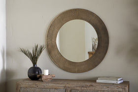 The nkuku Moto Round Mirror in Antique Brass hangs above a rustic wooden console table. On the table are a dark vase with tall green leaves, a white candle on a wooden tray, and two stacked books. The mirror reflects minimalist decor.