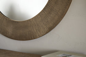 A close-up of the nkuku Moto Round Mirror in Antique Brass, featuring a textured frame and hanging on a light, subtly textured wall. A wooden surface with a closed book or box appears partially in the lower right corner.