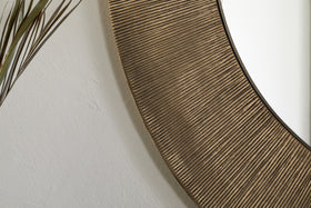 The Moto Round Mirror - Antique Brass by nkuku features a textured, grooved frame. In this close-up, it reflects a plain white wall, with thin brown plant leaves accenting the left edge for a minimalist look.