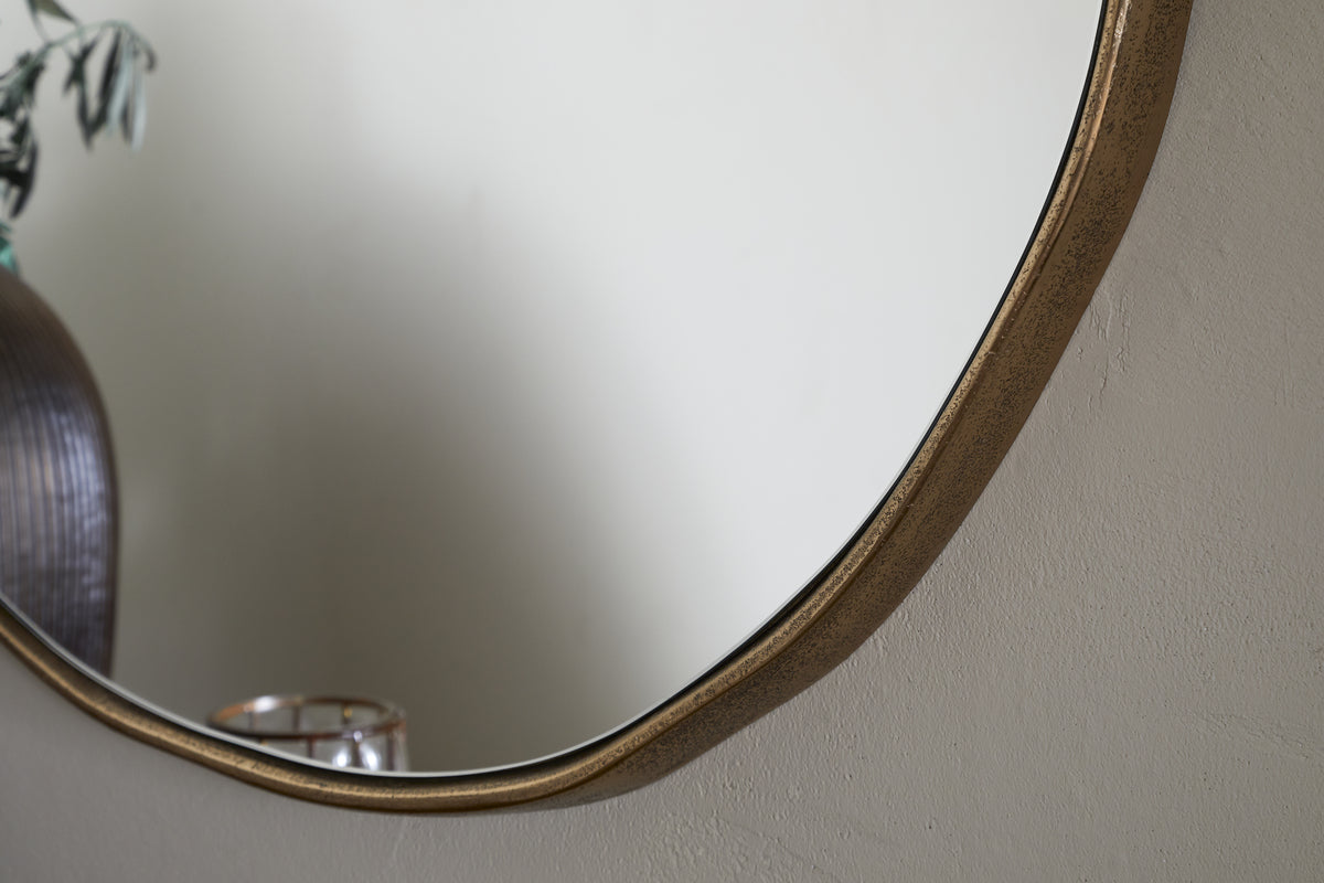 A close-up of the nkuku Mchanga Organic Shape Mirror in Antique Brass, hung on a light-textured wall. Its reflection shows part of a ribbed vase with greenery and a grid-pattern candle holder, both softly blurred.