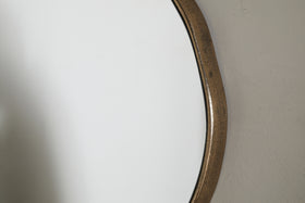Close-up of the nkuku Mchanga Organic Shape Mirror in antique brass, mounted on a textured, light wall. The frame has a slightly weathered look, and the round mirror reflects a bright, neutral background.