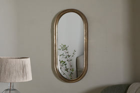 The Mbwa Oval Mirror in Antique Brass by nkuku hangs on a light gray wall. A tall lamp with a beige pleated shade stands to the left, while a light green sofa is partly visible on the right. The mirror reflects a green leafy plant.