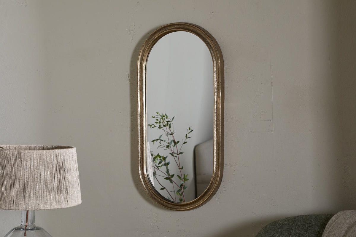 The Mbwa Oval Mirror in Antique Brass by nkuku hangs on a light gray wall. A tall lamp with a beige pleated shade stands to the left, while a light green sofa is partly visible on the right. The mirror reflects a green leafy plant.