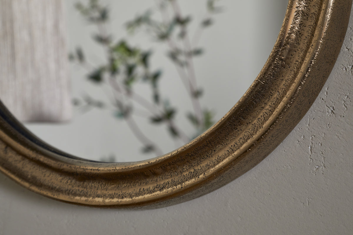 Close-up of the nkuku Mbwa Oval Mirror in antique brass, featuring an aged gold-toned frame on a light textured wall, reflecting leafy greenery and a beige curtain for a calm, relaxed ambiance.