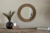 The Putali Etched Mirror - Antique Brass by nkuku hangs on a beige wall above a rustic wood console table topped with a black vase of dried grass, a white candle on wood, and stacked books for a minimal, stylish scene.