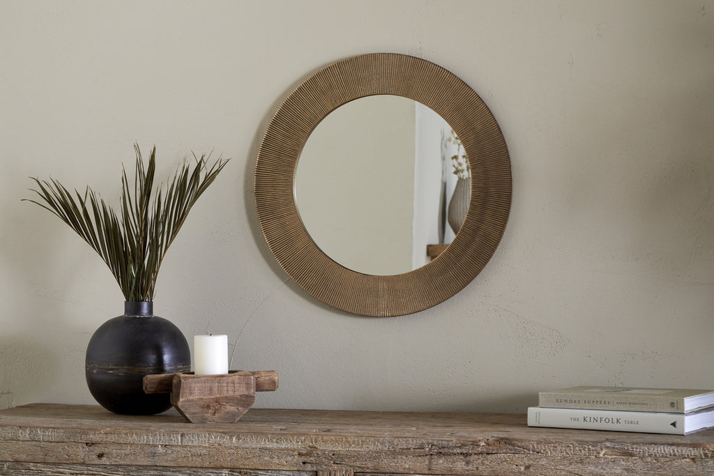 The Putali Etched Mirror - Antique Brass by nkuku hangs on a beige wall above a rustic wood console table topped with a black vase of dried grass, a white candle on wood, and stacked books for a minimal, stylish scene.
