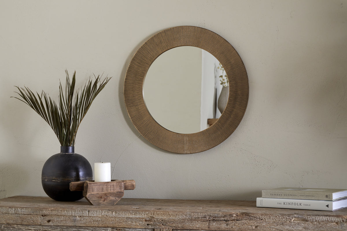 The Putali Etched Mirror - Antique Brass by nkuku hangs on a beige wall above a rustic wood console table topped with a black vase of dried grass, a white candle on wood, and stacked books for a minimal, stylish scene.