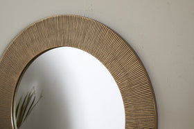 Close-up of the Putali Etched Mirror - Antique Brass by nkuku, showcasing its textured gold-toned frame with fine vertical grooves. The round mirror reflects thin green plant leaves against a plain off-white wall.