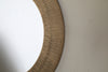 Close-up of the Putali Etched Mirror - Antique Brass by nkuku, featuring a sculptural, wide wooden frame with vertical grooves. The round mirror hangs on a light wall, reflecting a clean white space.