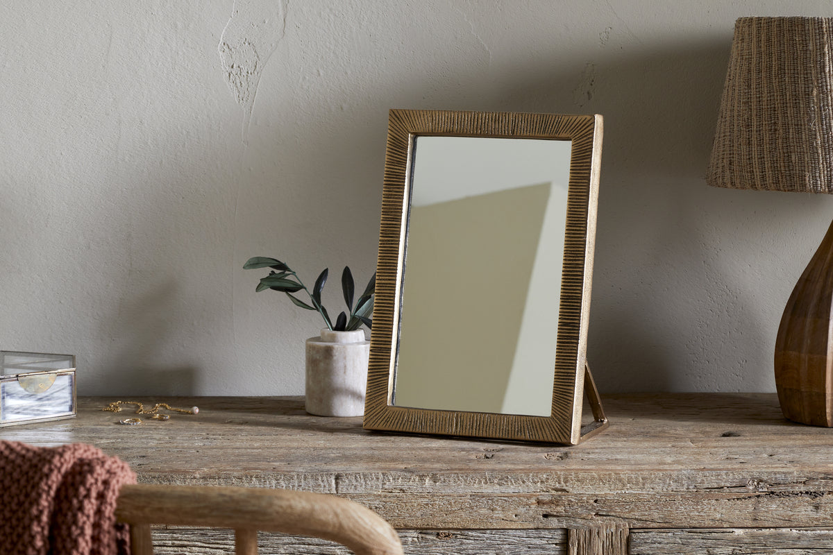 The nkuku Baja Standing Mirror - Antique Brass stands on a rustic wooden table beside a potted plant, a jewelry box with scattered jewelry, and a brown lamp with woven shade. A wooden chair and knitted blanket are in the foreground.