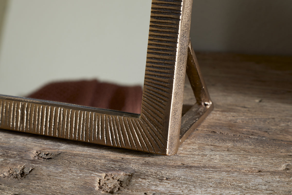 A close-up of the nkuku Baja Standing Mirror in antique brass, featuring a textured metal frame. The mirror rests on a rustic wooden surface with visible grain, and its support stand is partially visible behind the frame.
