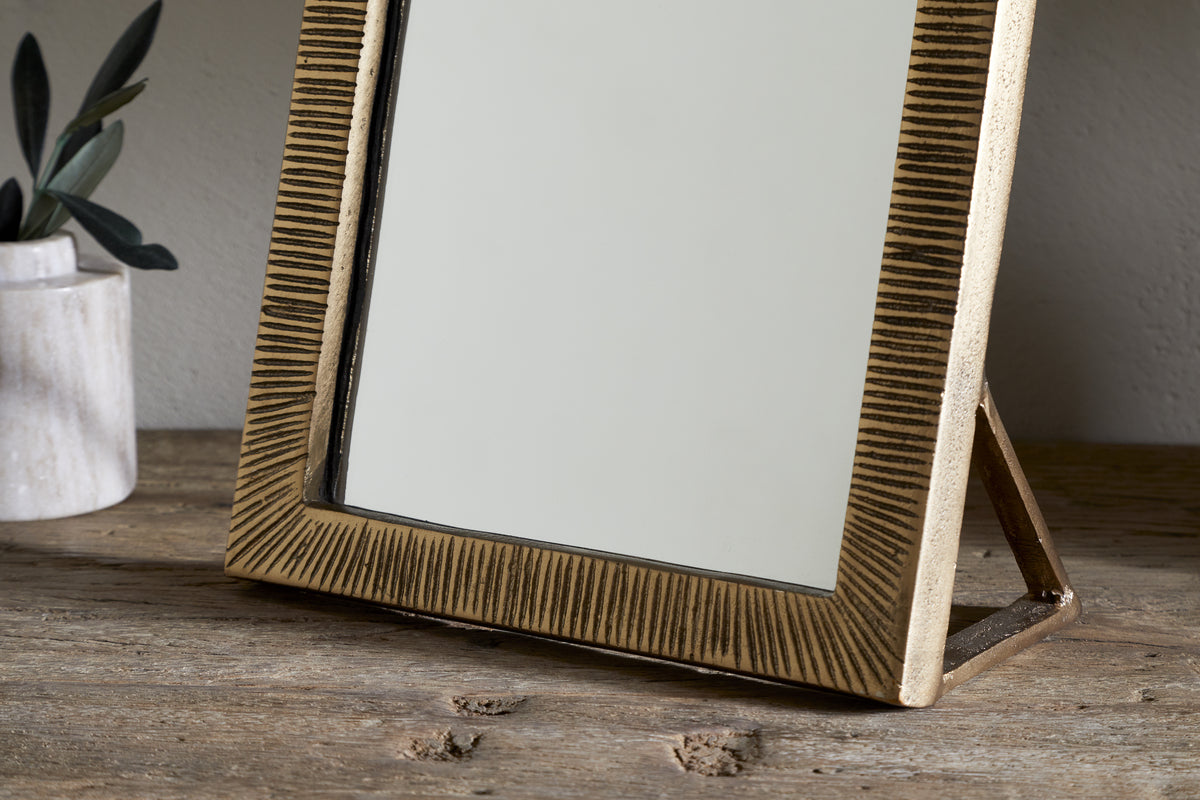 The nkuku Baja Standing Mirror in Antique Brass with a textured frame and stand is displayed on a rustic wooden surface, accompanied by a small marble vase with green stems to the left in a softly lit, minimal setting.