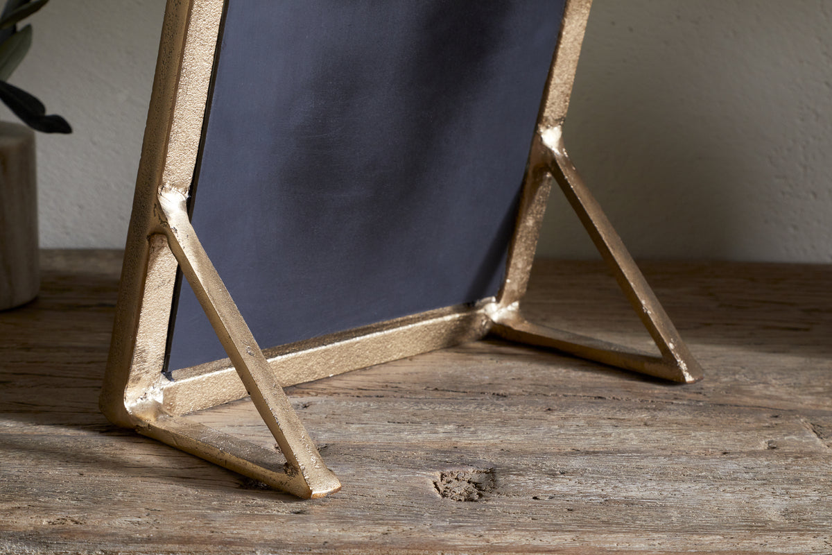 A close-up of the nkuku Baja Standing Mirror in antique brass, showing its rectangular frame and angled metal supports on a rustic wooden surface. A gray ceramic pot with green leaves appears in the background to the left.