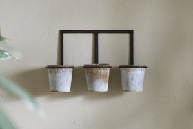 Three zinc pots from the Tukana Triple Wall Hung Planter by nkuku hang from a black wall bracket against a beige textured wall. The pots are empty, with green leaves partly visible in the left foreground.