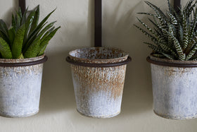 The nkuku Tukana Triple Wall Hung Planter - Zinc features three weathered metal pots; spiky green succulents fill the left and right, while the distressed middle planter is empty with visible rust marks.