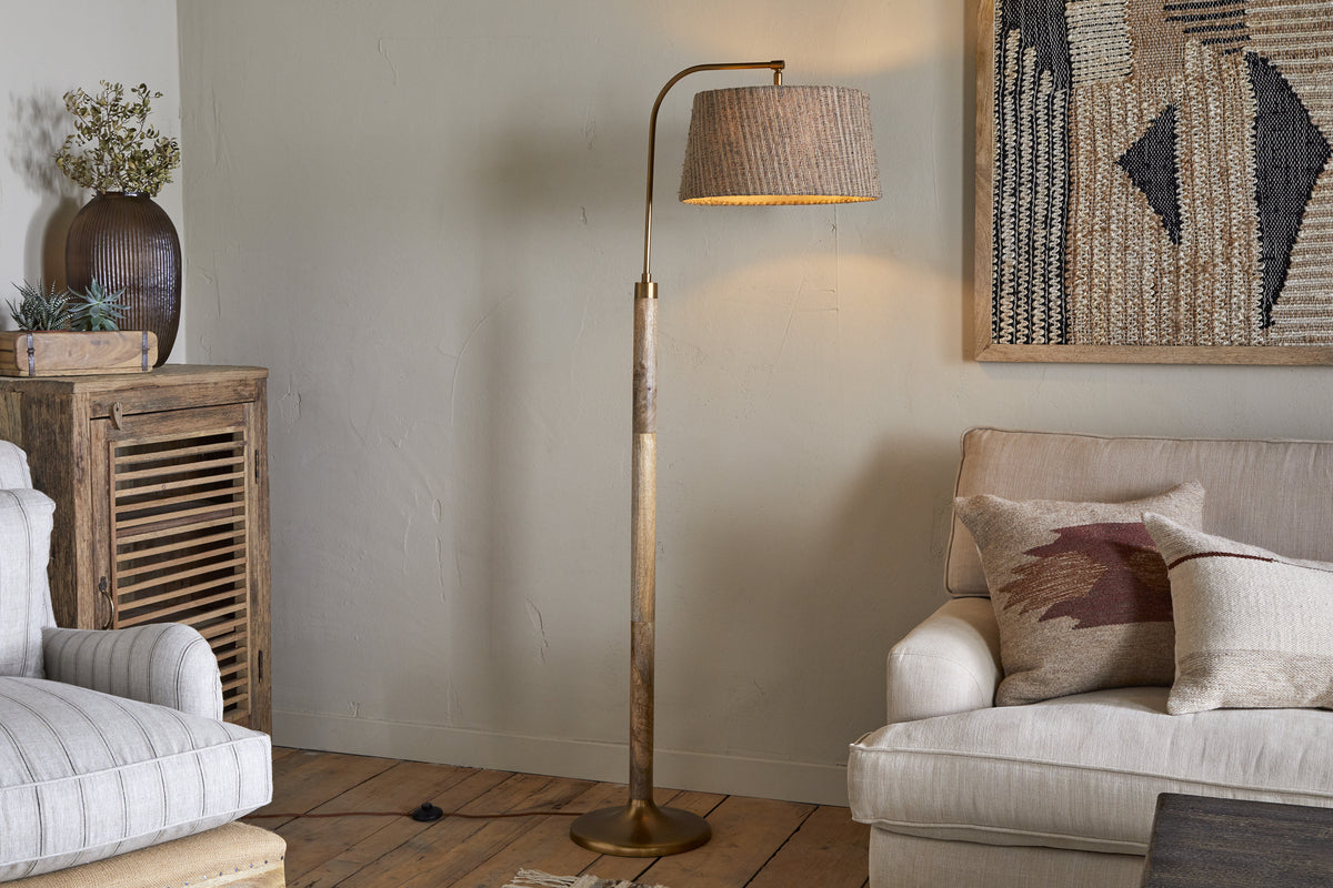 A cozy living room with the nkuku Kera Wood & Metal Floor Lamp in antique brass and natural beside a light sofa with patterned cushions, near a rustic mango wood base cabinet, abstract wall art, and a potted plant on the cabinet.