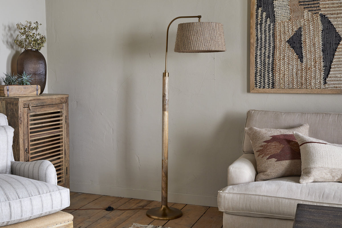 A cozy living room with a beige armchair, cream sofa with pillows, wooden furniture, plants, and a textured wall hanging. The space features the nkuku Kera Wood & Metal Floor Lamp in antique brass & natural mango wood.