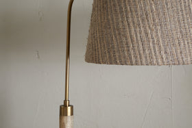 Close-up of the nkuku Kera Wood & Metal Floor Lamp in Antique Brass & Natural, showing part of its brass arm and textured beige shade against a light-colored, cracked wall.
