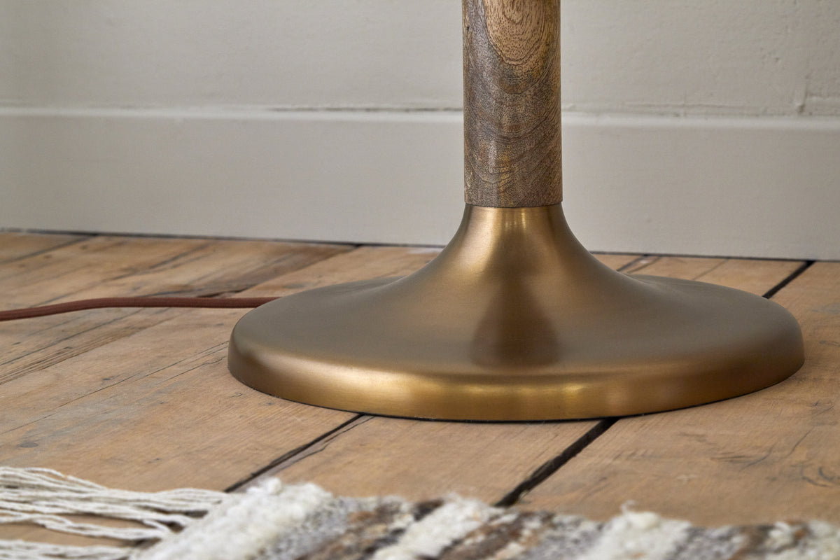 A close-up of the nkuku Kera Wood & Metal Floor Lamp in Antique Brass & Natural shows its round mango wood base and wooden pole on a wooden floor, with a brown cord trailing near a woven rug with white tassels against a light wall.