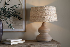The Dalat Ceramic Table Lamp - Natural by nkuku, featuring a curvy base and woven beige shade, sits on a rustic console table with books and a leafy branch in a dark vase. Neutral art decorates the textured beige wall behind.