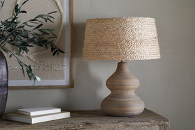A wooden table displays the Dalat Ceramic Table Lamp - Natural by nkuku, featuring a wide, woven straw shade. Beside it are stacked books and a dark vase with green branches. Beige textured wall art decorates the light-colored wall behind.