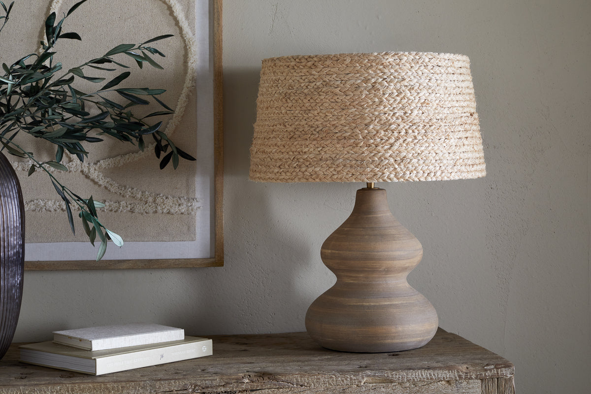 A wooden table displays the Dalat Ceramic Table Lamp - Natural by nkuku, featuring a wide, woven straw shade. Beside it are stacked books and a dark vase with green branches. Beige textured wall art decorates the light-colored wall behind.