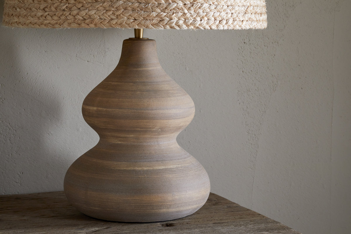 A close-up of the Dalat Ceramic Table Lamp - Natural by nkuku, featuring a curvy matte brown ceramic base and a woven natural fiber shade, set on a rustic wooden surface against a light gray textured wall.