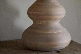 The Dalat Ceramic Table Lamp - Natural by nkuku features a smooth, light brown base with curvy, hourglass lines. Set on rustic wood against a neutral wall, its handcrafted look and subtle stripes are beautifully highlighted by natural light.