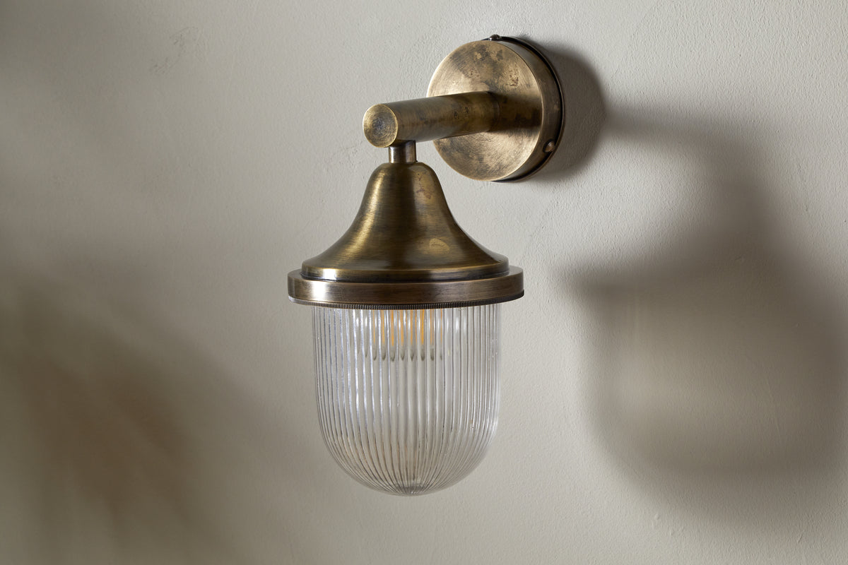 The nkuku Magola Outdoor Wall Lamp in antique brass is mounted on a light wall, featuring a rounded metal top and clear, vertically ribbed glass that casts soft shadows and warm ambient light.