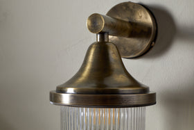 The nkuku Magola Outdoor Wall Lamp in antique brass features a round backplate, conical shade, and ribbed clear glass, casting a warm ambient glow on exterior walls.