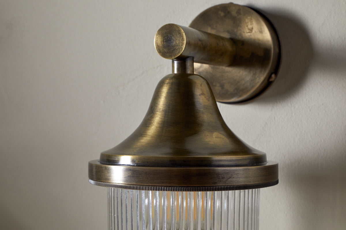 The nkuku Magola Outdoor Wall Lamp in antique brass features a round backplate, conical shade, and ribbed clear glass, casting a warm ambient glow on exterior walls.